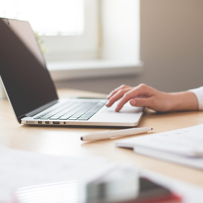 Eine Hand tippt auf einem Laptop, im Hintergrund sind Notizen und ein Smartphone zu sehen.